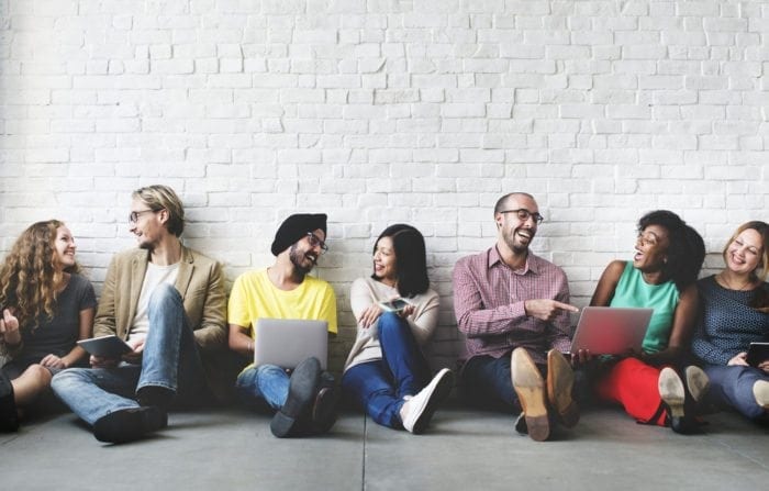 young people sit leaning against a wall and using laptop
